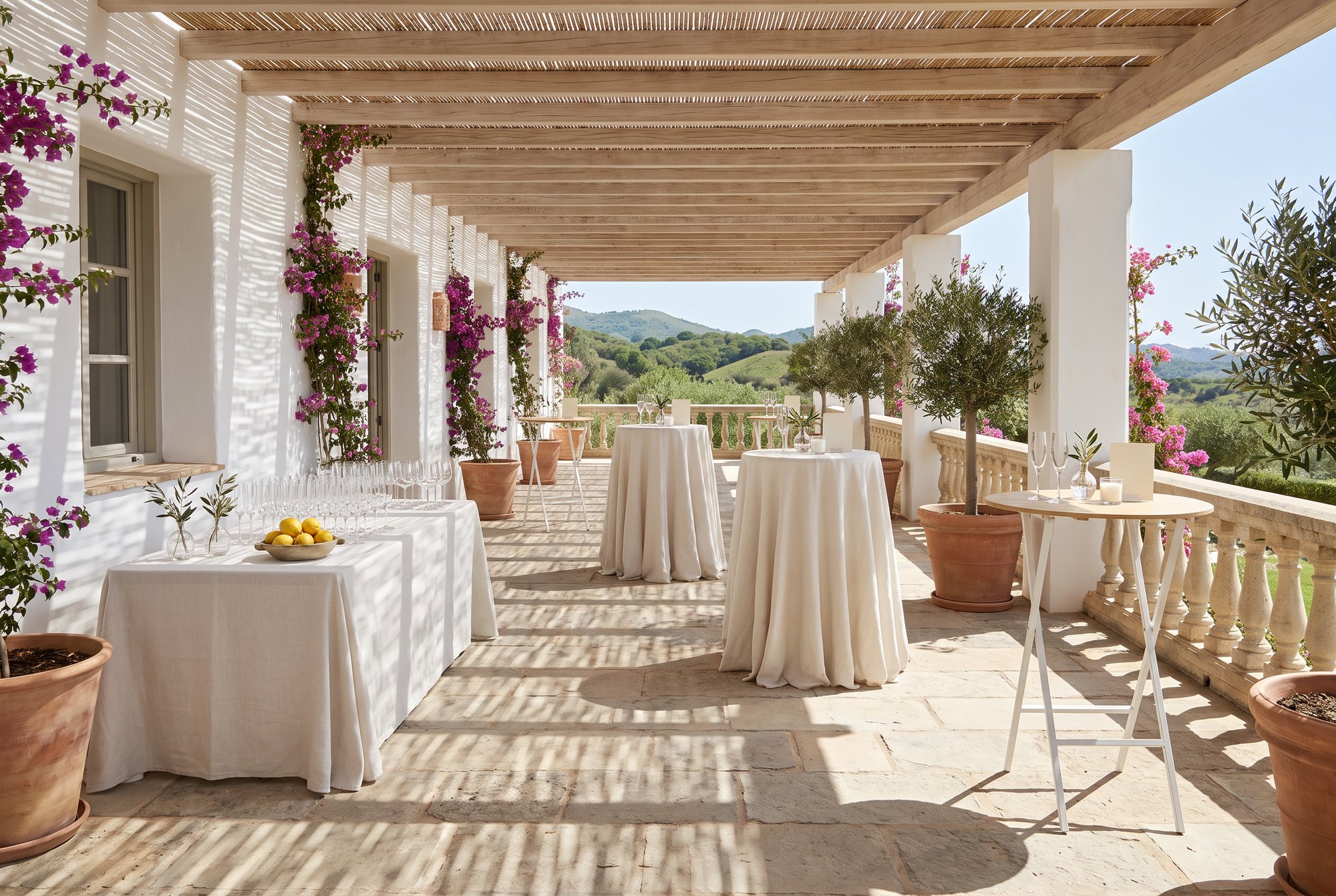 Terraza de un cortijo bajo una pérgola: dos mesas altas con mantel de lino, otras sin mantel, y una mesa larga de bienvenida con copas y limones.