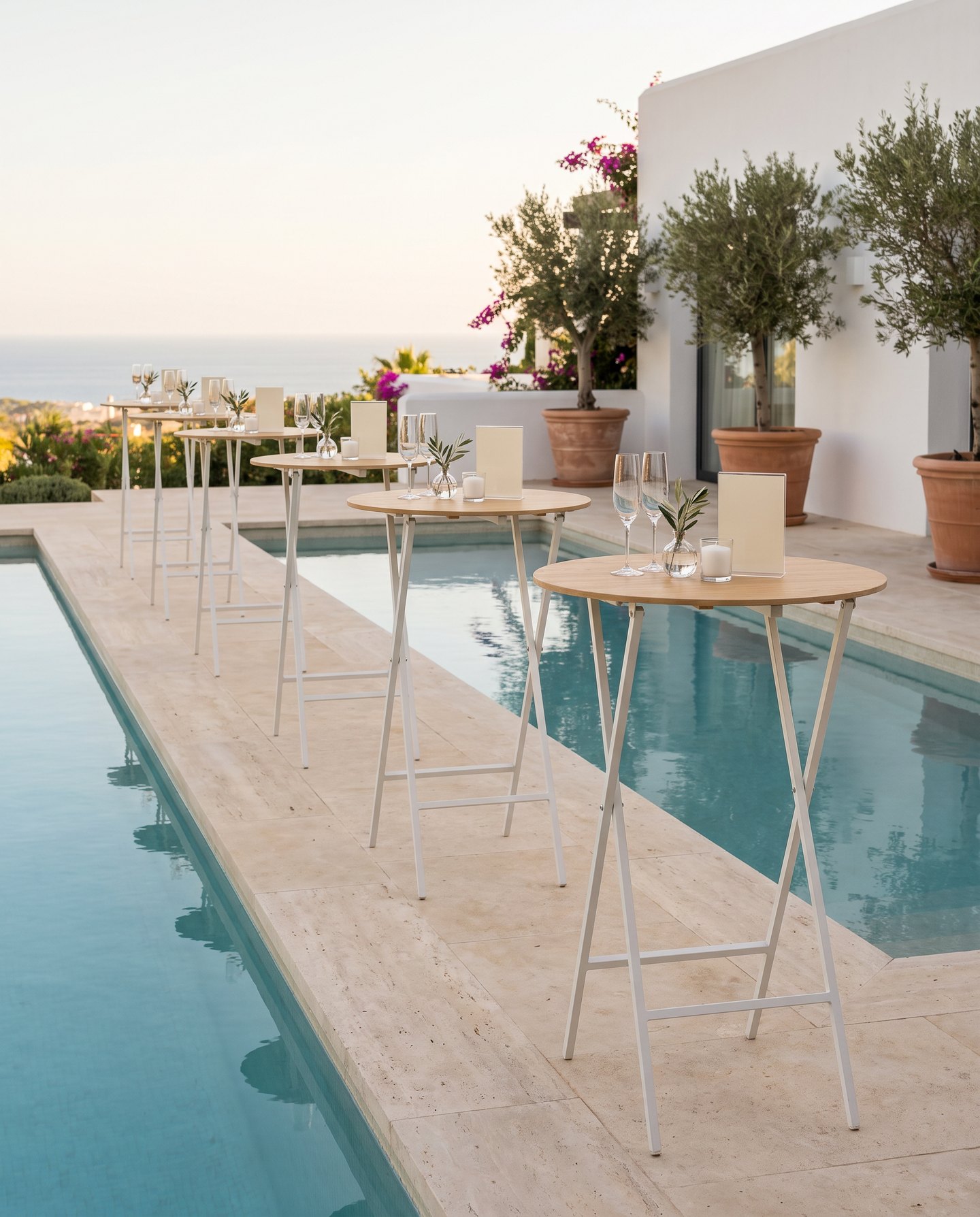 Mesas altas en fila junto a la piscina de una villa en Marbella, cada una con copas de cava, una rama de olivo y una vela.